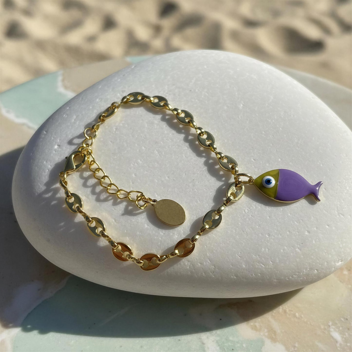 Gold bracelet with a fish charm on a white stone with sand background