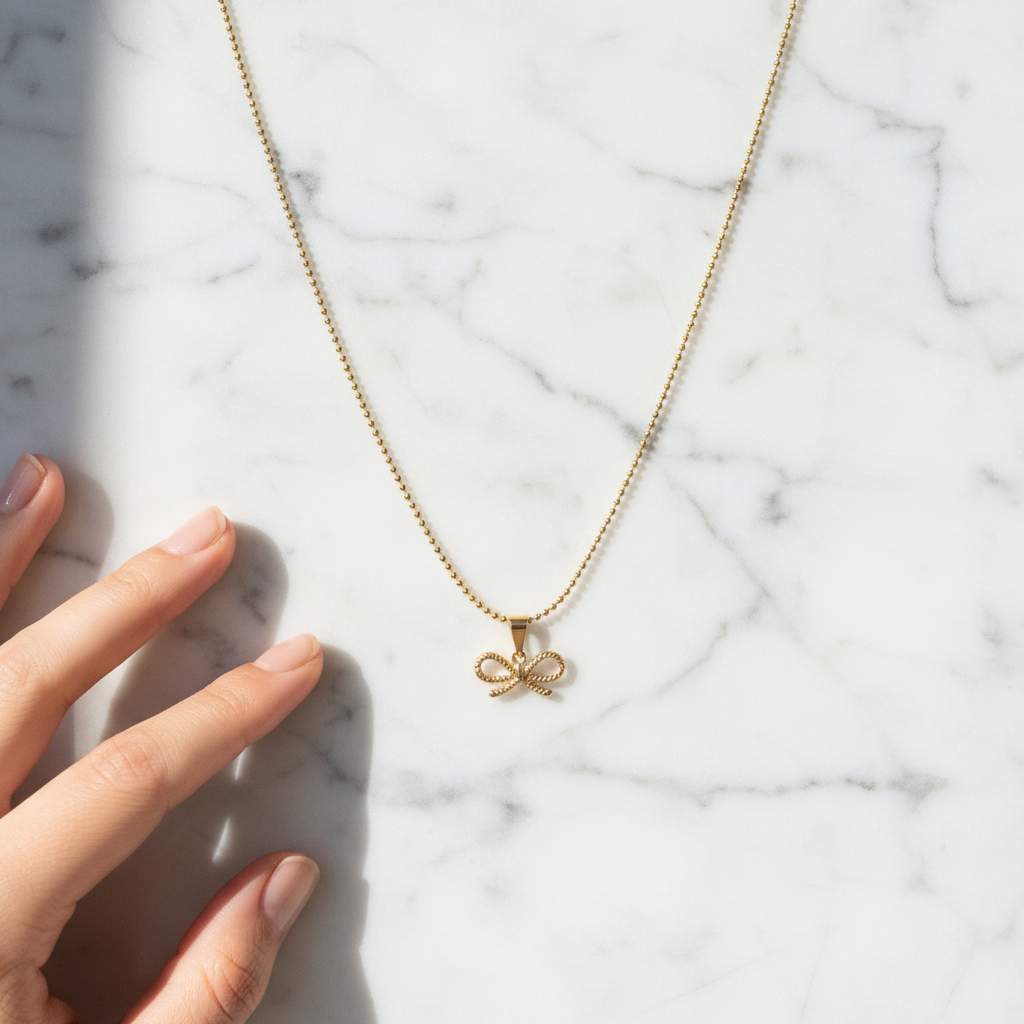 Gold Chica Chica Jewelry necklace with a bow pendant on a marble surface next to a hand.