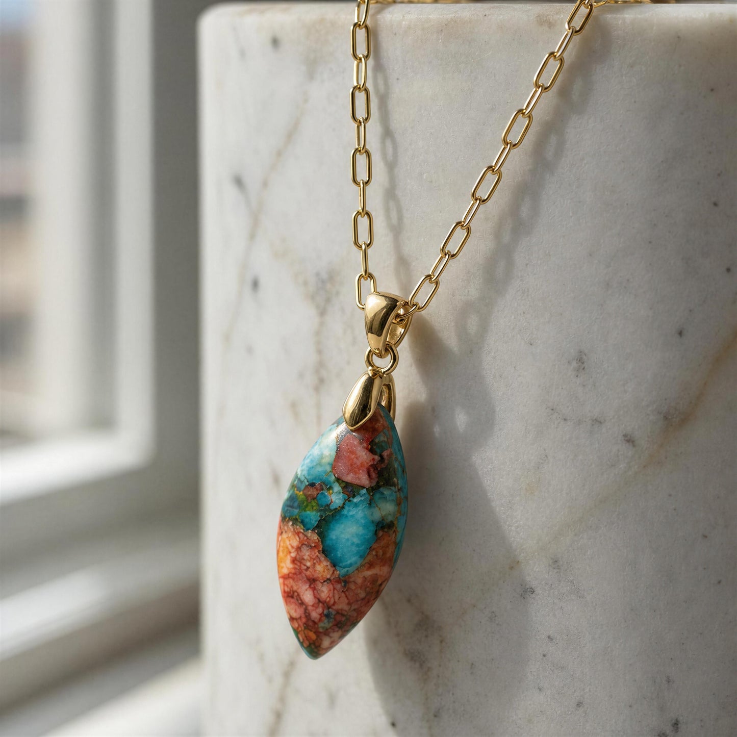 The Pietra Alba Natural Stone Necklace from Chica Chica Jewelry,  a gold paperclip chain 18K gold plated necklace with a marquise-shaped pendant featuring a vibrant turquoise, orange, and brown marbled pattern, resting on a white marble cylinder.
