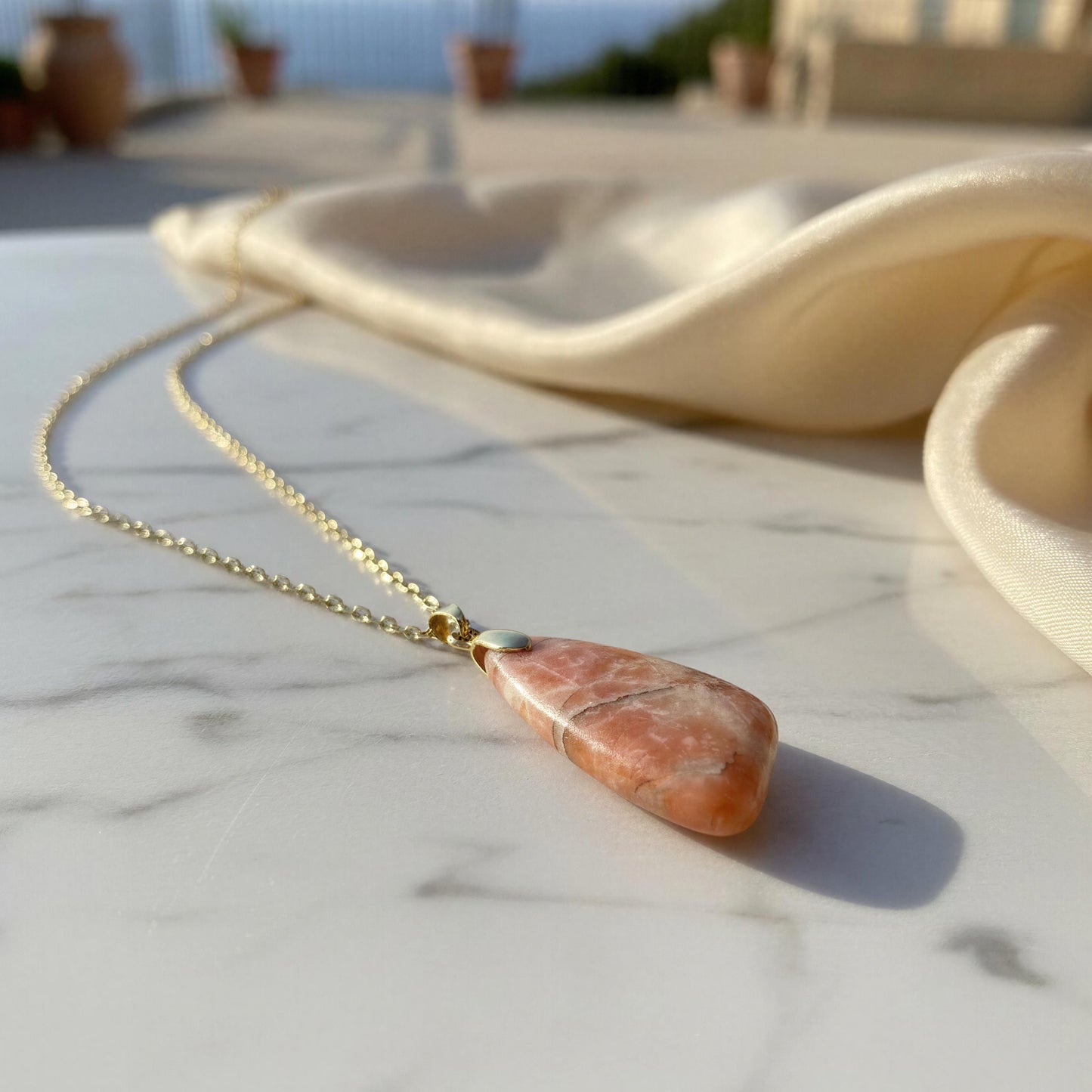 A gold chain necklace with a peach-orange stone pendant featuring white veining, resting on a white marble surface with a cream satin cloth and a blurred outdoor background.