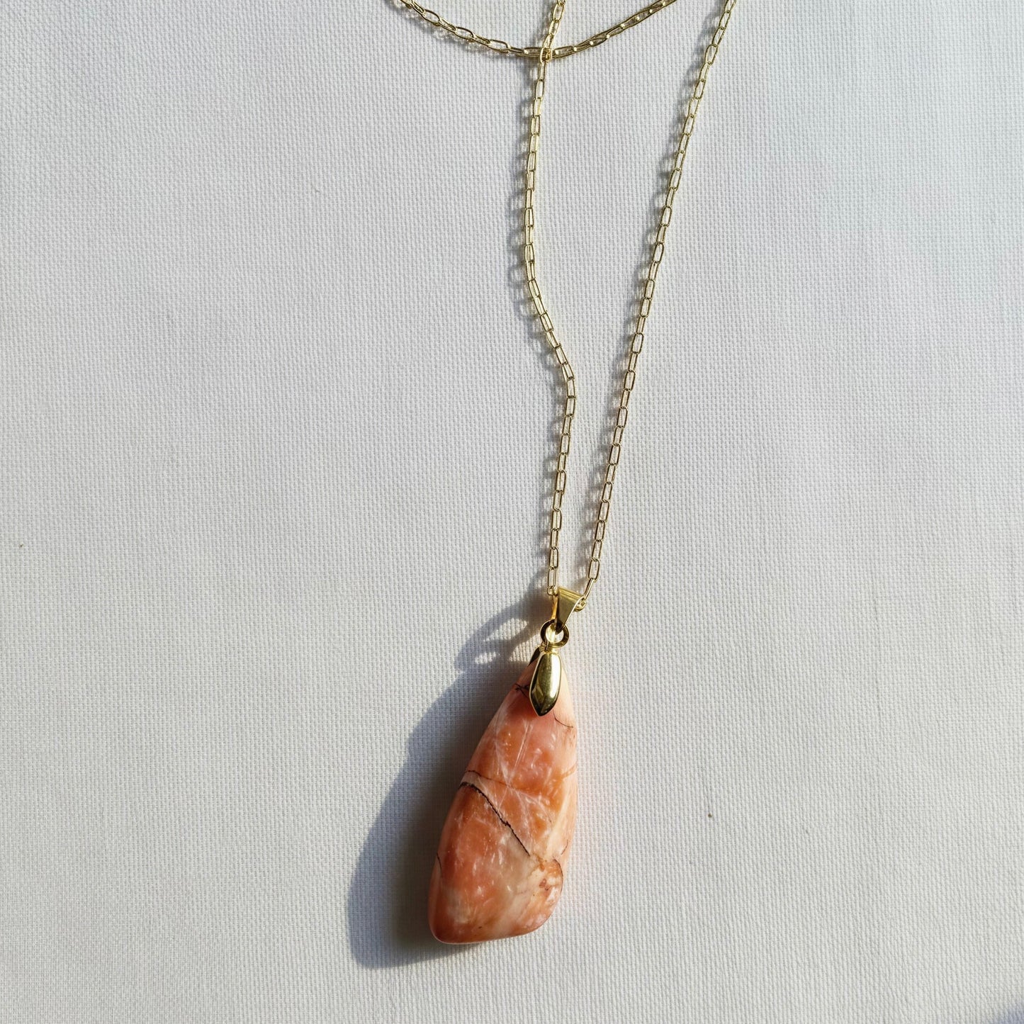 A gold chain necklace featuring a teardrop-shaped peach and orange stone pendant with natural fissures, displayed on a textured white background.