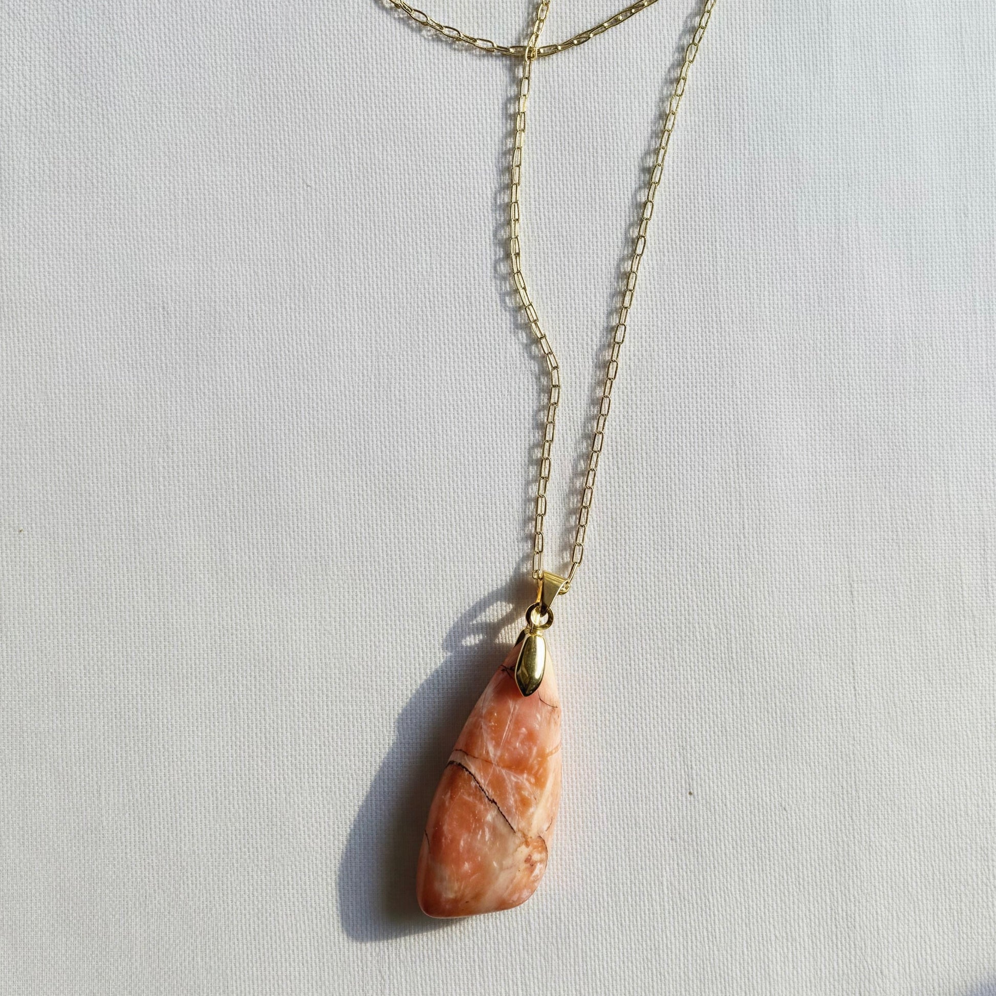 A gold chain necklace featuring a teardrop-shaped peach and orange stone pendant with natural fissures, displayed on a textured white background.