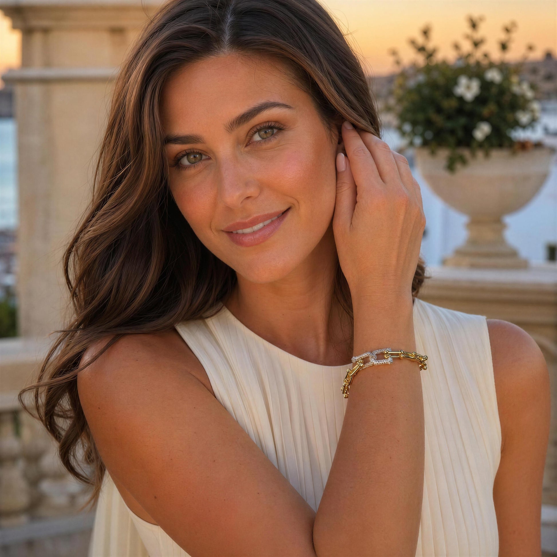 A woman with long brown hair wearing a cream pleated sleeveless top and the Radiance Link Bracelet with 18K gold-plated links and silver-tone pavé zircon centerpiece, posing outdoors with a warm sunset background.