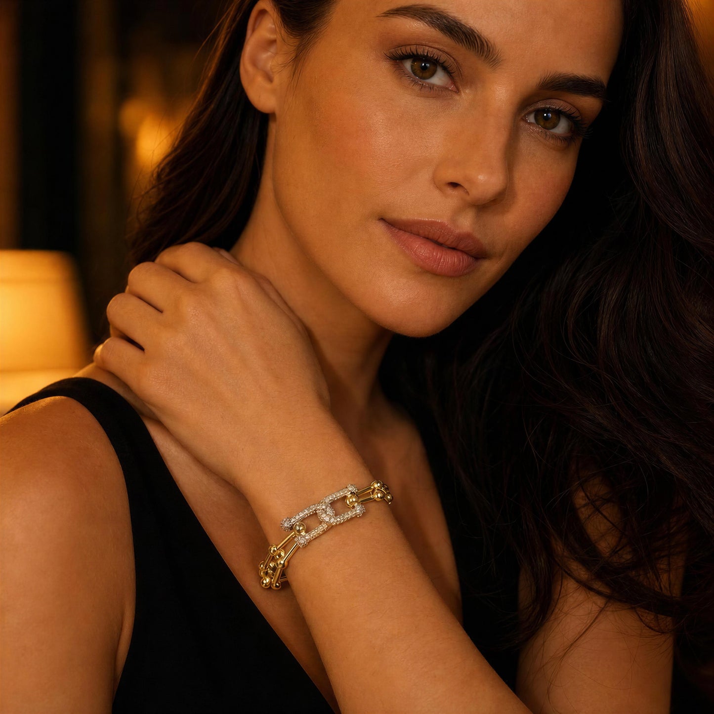 A close-up lifestyle shot of a woman wearing a gold chain bracelet with clear pavé-set stones on her wrist, set against a warm, softly blurred background.