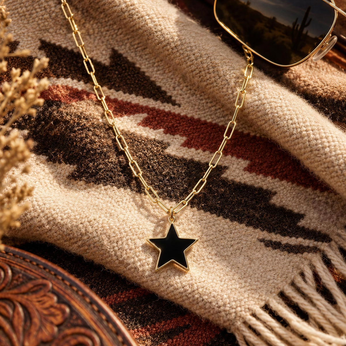 The Radiant Noir Star Necklace from Chica Chica Jewelry, a gold chain necklace with a black star pendant, styled on a southwestern patterned blanket with sunglasses and dried flowers.