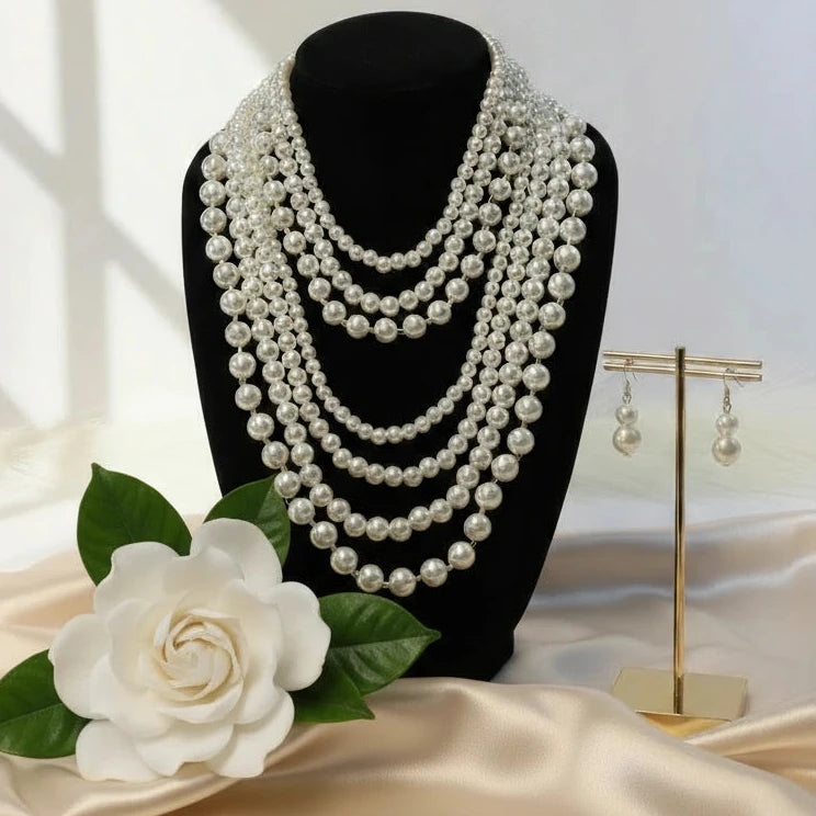 Multiple layers of pearl necklace on a black mannequin with a white flower and earring stand in the background.