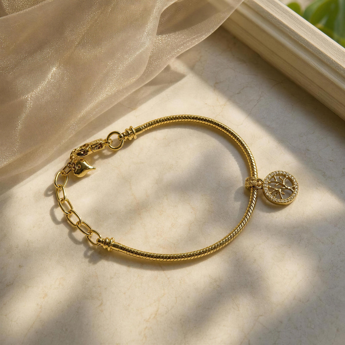 The Renata gold Tree of Life charm bracelet by Chica Chica Jewelry, displayed on a light marble surface with soft beige fabric and natural shadows.