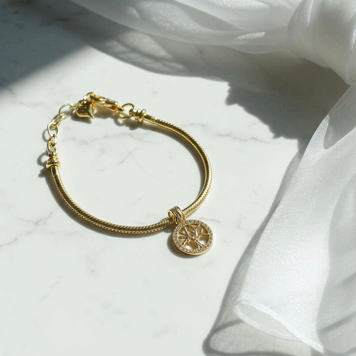 The Renata gold Tree of Life charm bracelet by Chica Chica Jewelry, displayed on a white marble surface with draped white fabric and soft sunlight shadows.