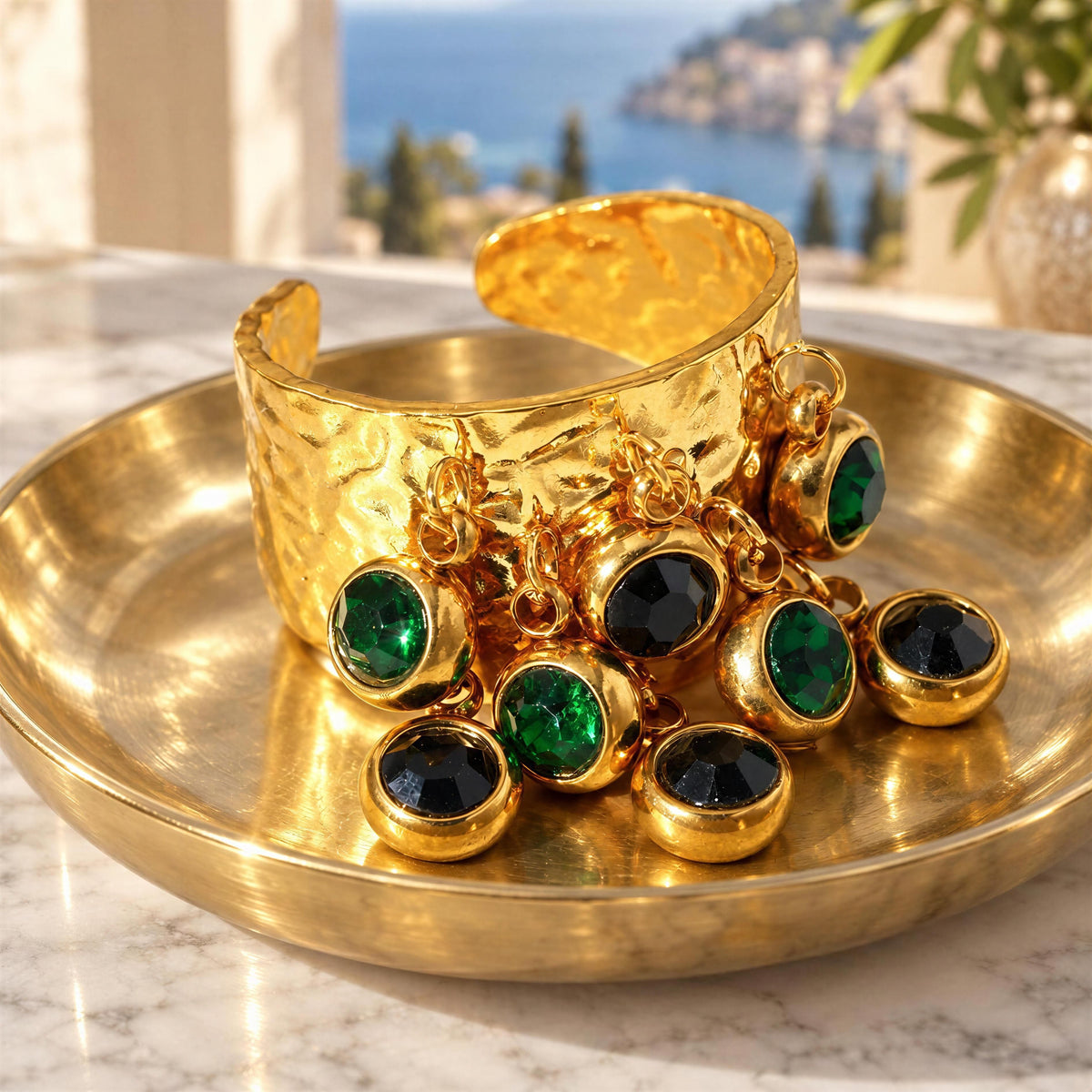 A chunky hammered gold ring with dangling gold chains and round green and black gemstone accents, displayed on a matching gold tray over a marble surface with a blurred coastal background.