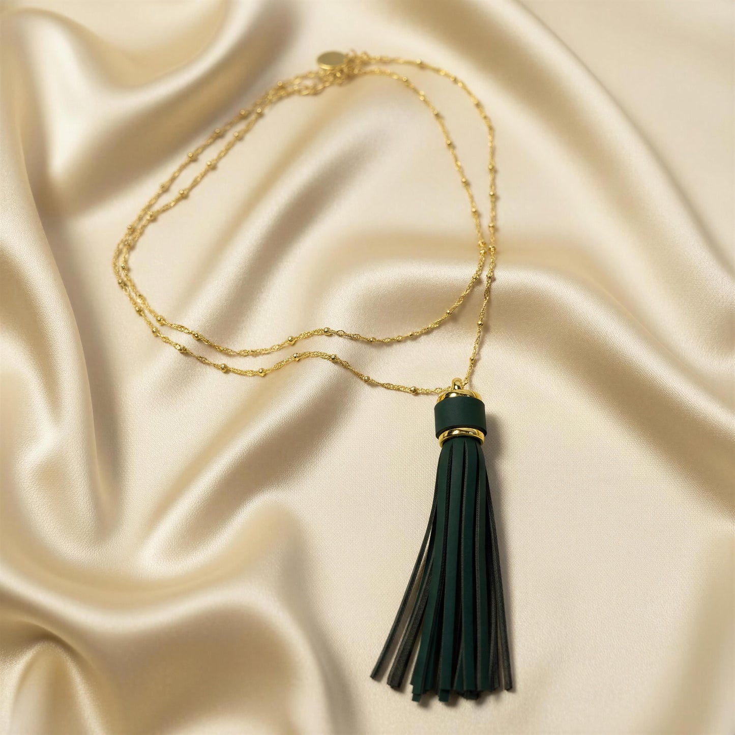 The Frangia Verde Necklace from Chica Chica Jewelry  featuring a dark green tassel pendant with a gold accent band, displayed on a draped cream satin fabric background.