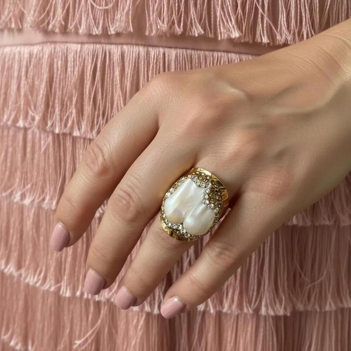 A close-up of a hand wearing a large gold ring with a white mother-of-pearl cabochon and small clear stones, set against a pink fringed garment background.