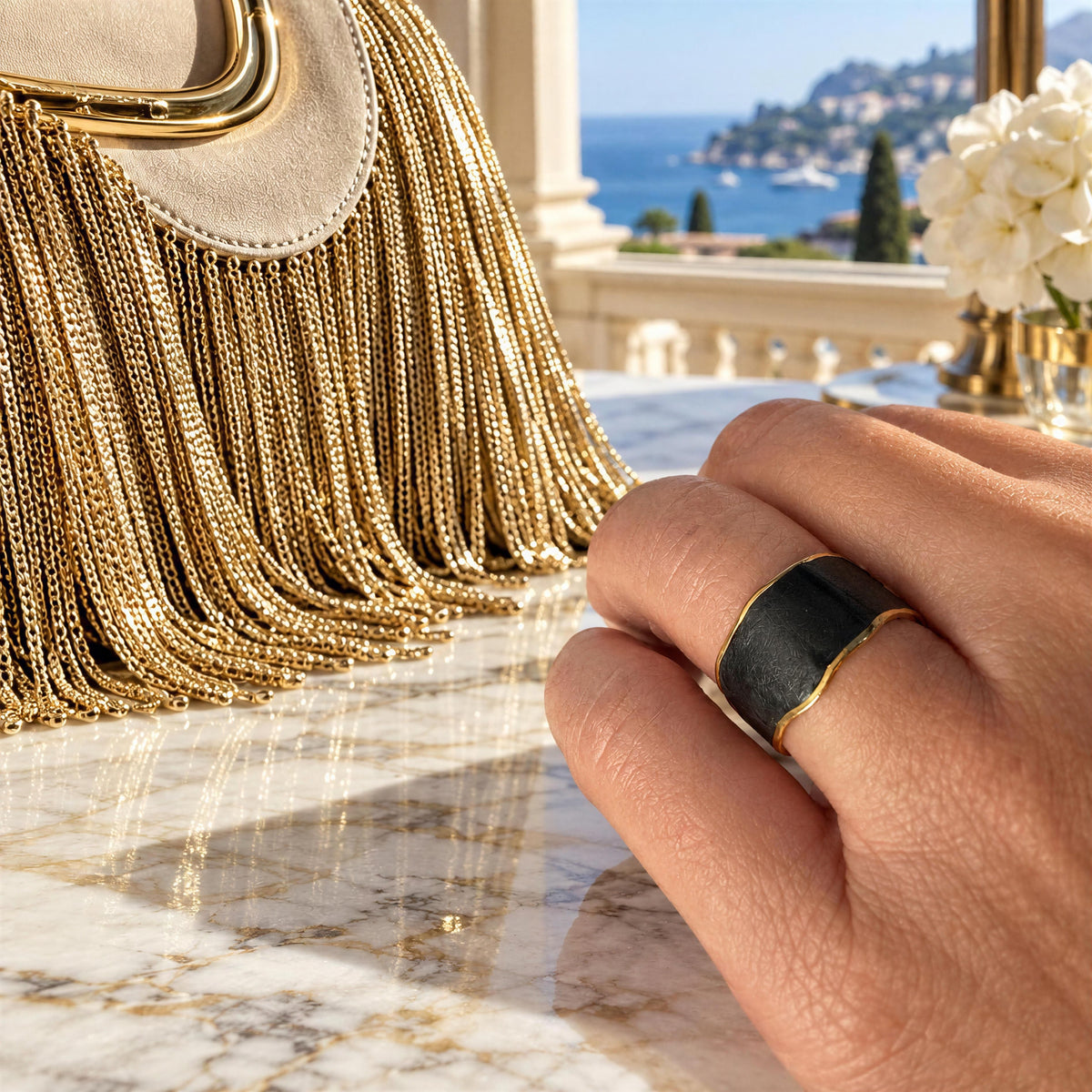 A close-up lifestyle shot of a hand wearing the Duna Ring – Ónice from Chica Chica Jewelry,  a wide black ring with gold trim, set against a luxurious background featuring a gold fringe handbag and a scenic coastal view.