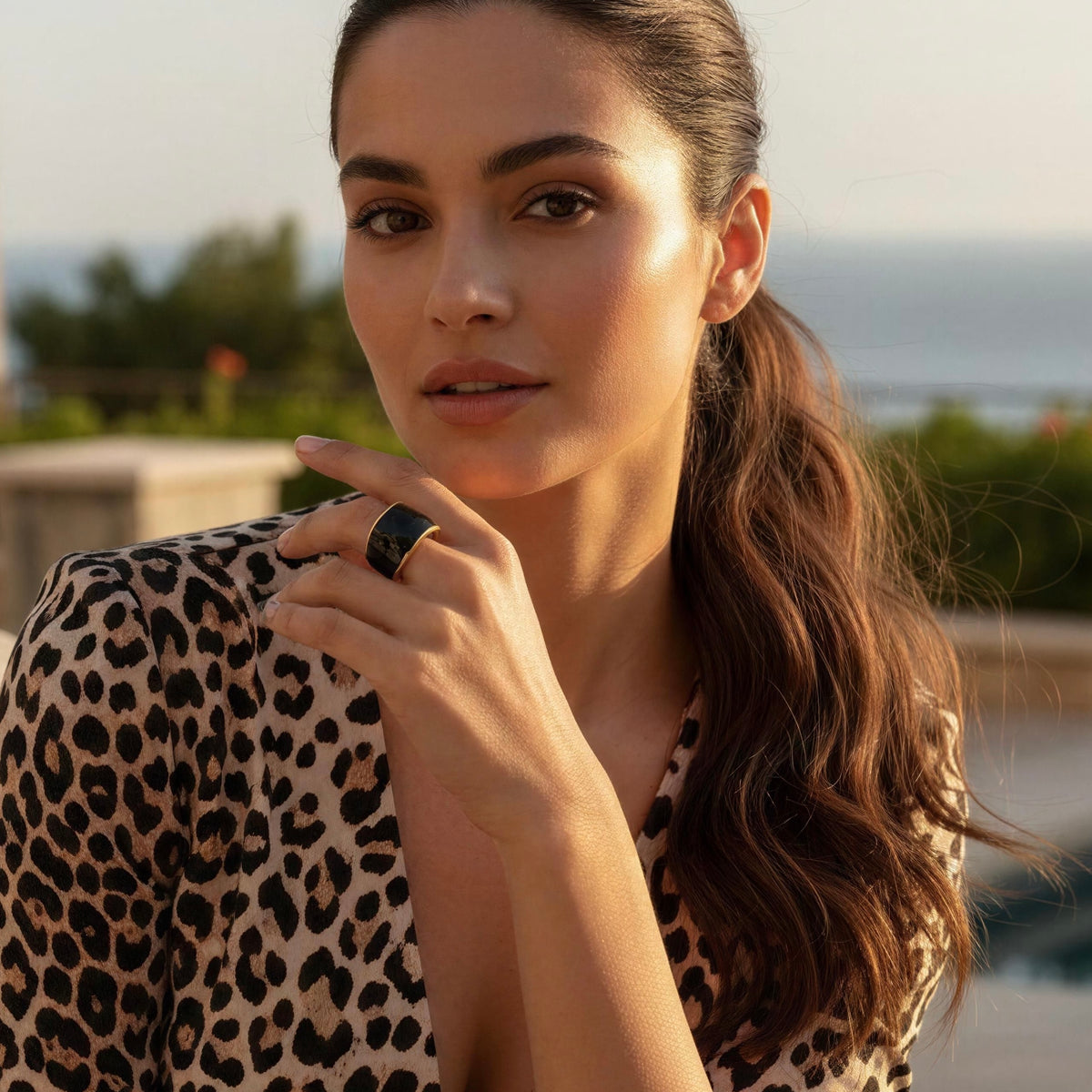 A woman with long brown hair tied back wears the  Duna Ring – Ónice from Chica Chica Jewelry  a black ring with a gold band on her finger, posing outdoors near a pool with a leopard-print top in the background.