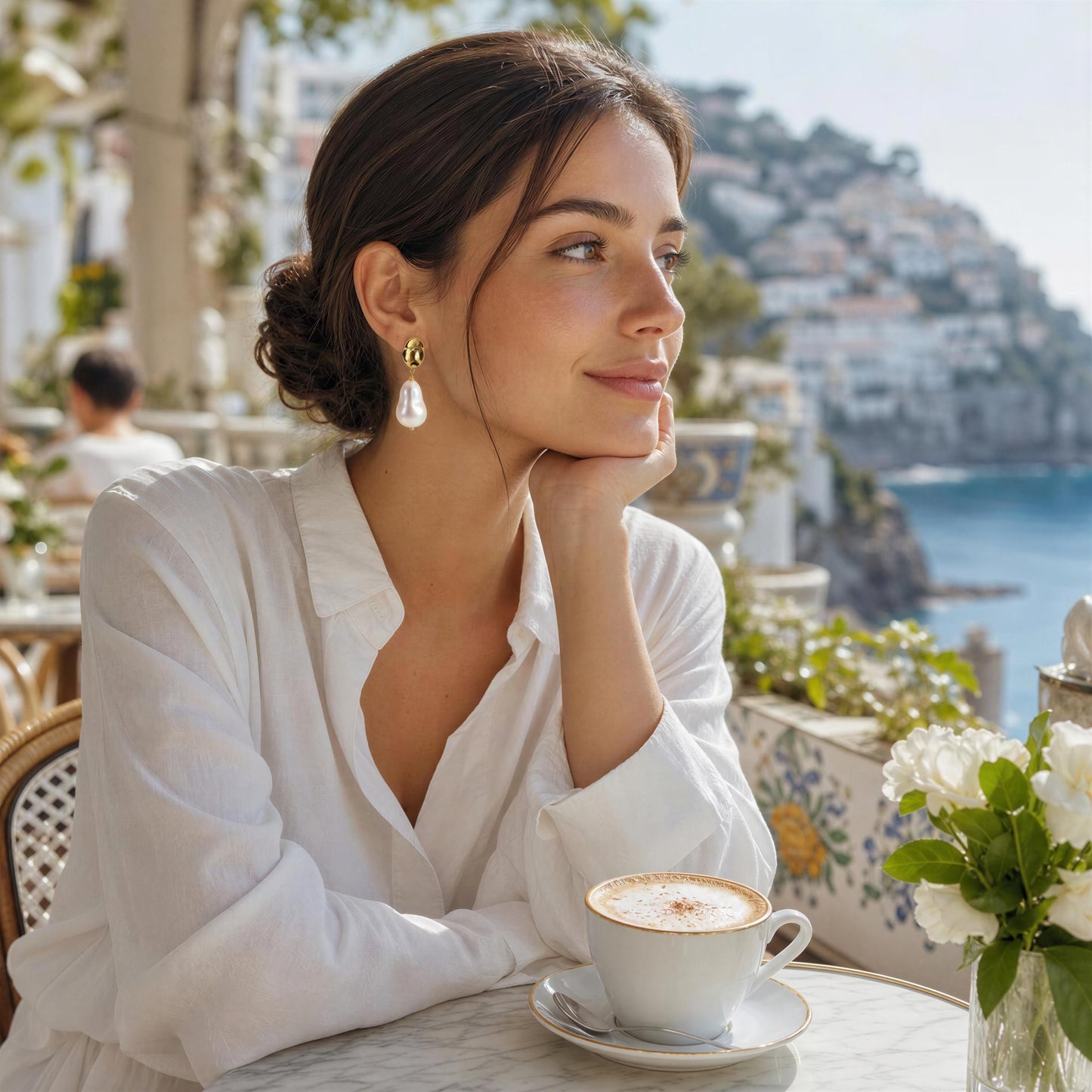 A woman in a white blouse sits at an outdoor table by the sea, wearing the Bellina Drop Pearl earrings. A cup of cappuccino and white flowers are on the table in front of her, with a coastal town in the background.