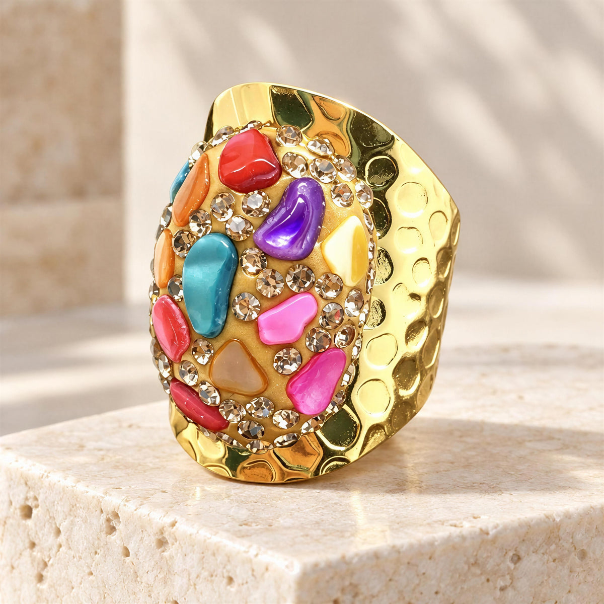 The Chroma Ring  from  Chica Chica Jewelry.  A chunky gold ring with a hammered texture, embellished with colorful faux-stone gems in pink, purple, blue, red, and orange, set on a beige stone pedestal.