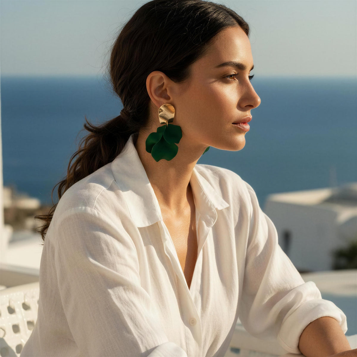 A woman in a white linen shirt wearing the Fiore Verde Earrings from Chica Chica Jewelry  , set against a sunny coastal background with the blue sea and white buildings.