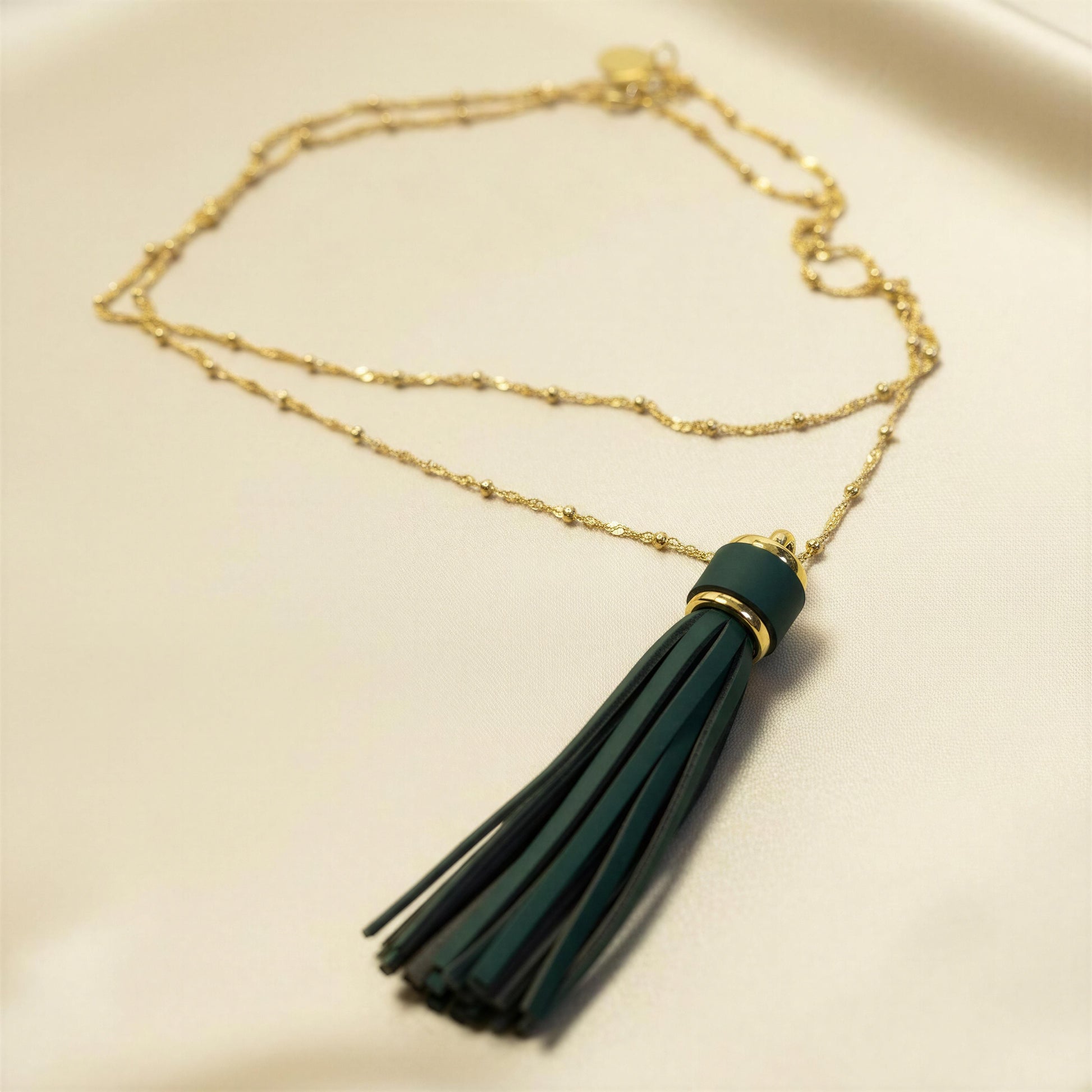 The Verde Necklace from Chica Chica Jewelry,  featuring a dark green tassel pendant with a gold accent ring, displayed on a light cream background.