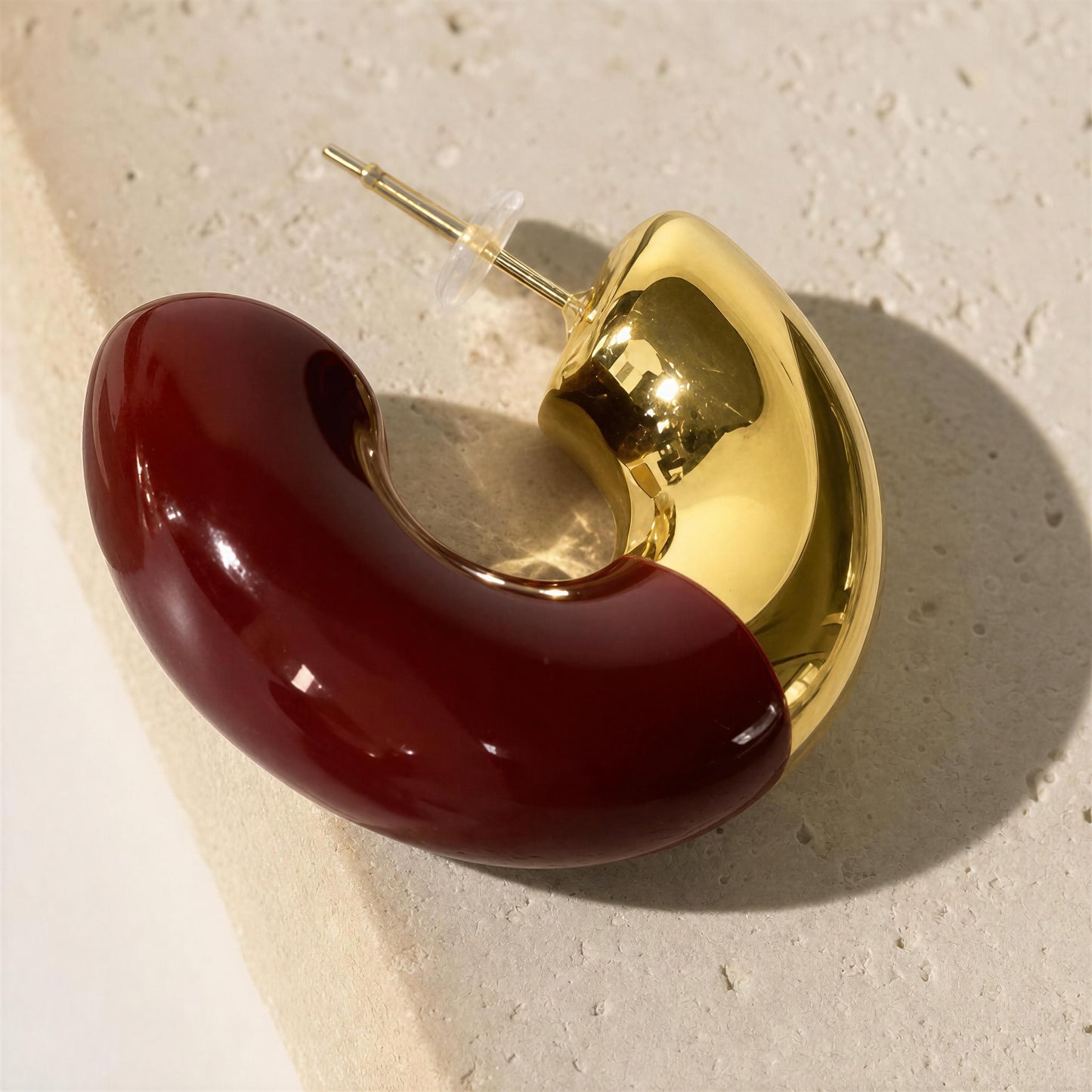 A glossy, curved two-tone earring with a deep burgundy red half and a polished gold half, shown on a textured beige stone surface.