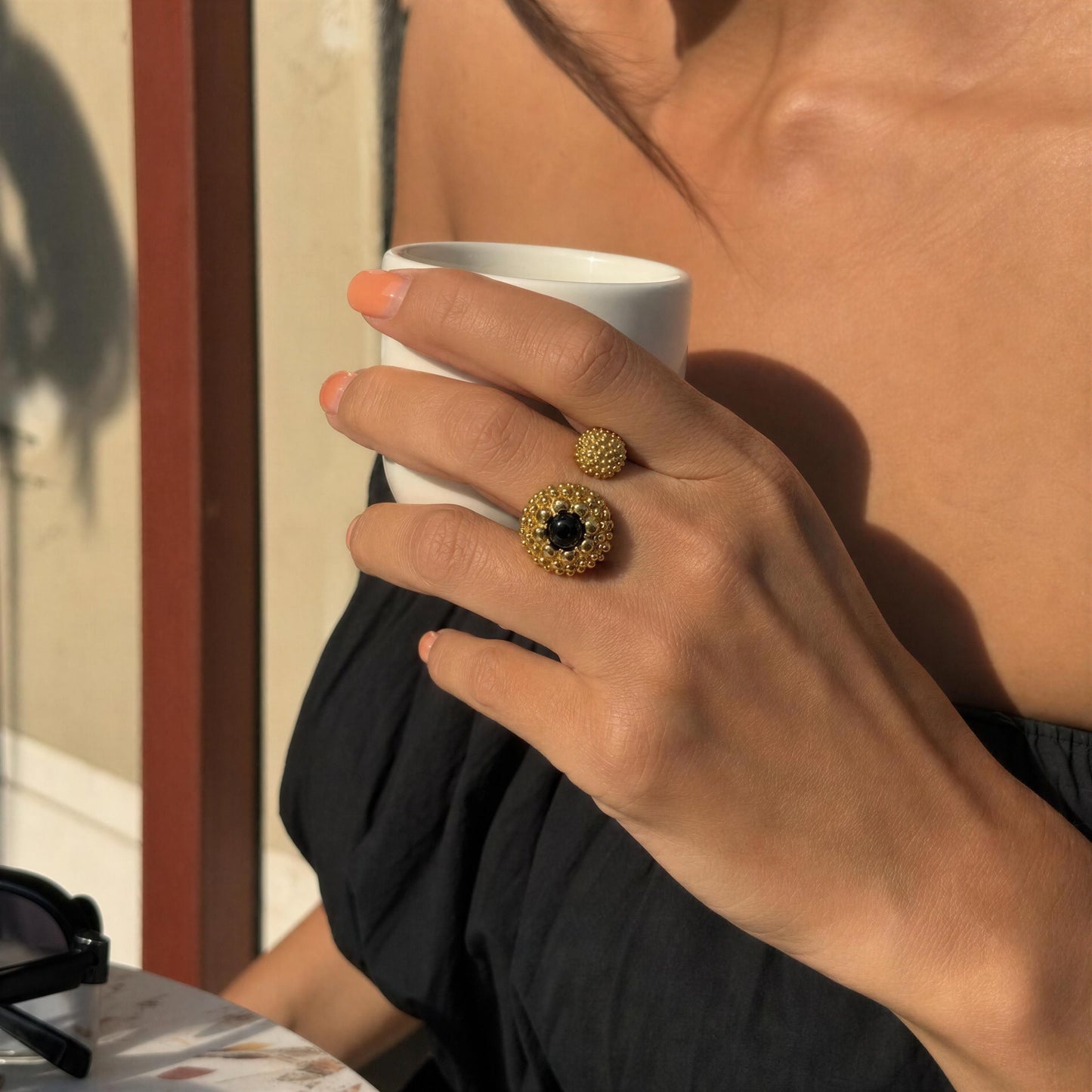 A close-up lifestyle shot of a person holding a white coffee cup, showcasing a textured gold ring with a black agate stone center.