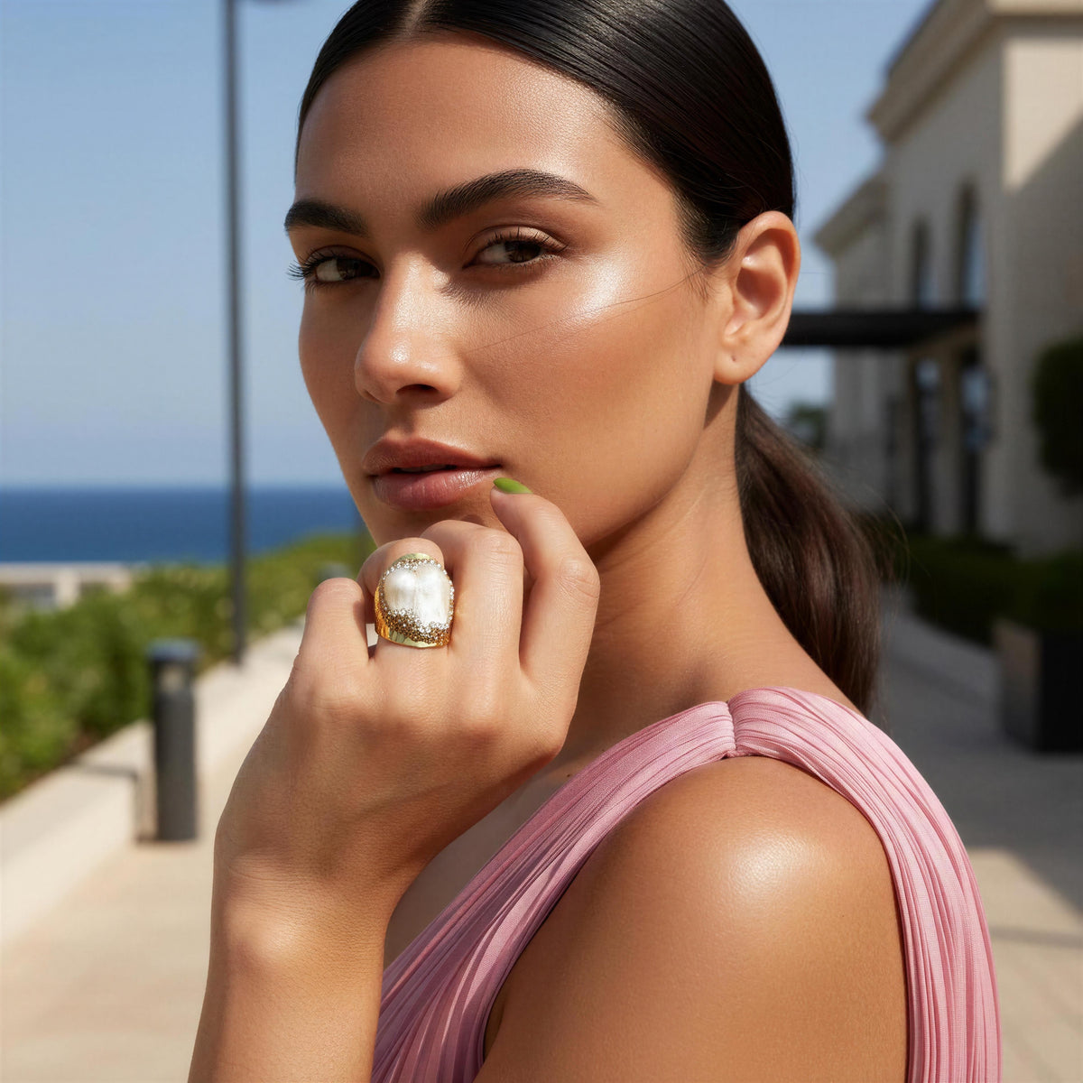 A woman in a pink pleated top wearing a large statement ring with a white stone and gold detailing, posing outdoors near a beach and building.