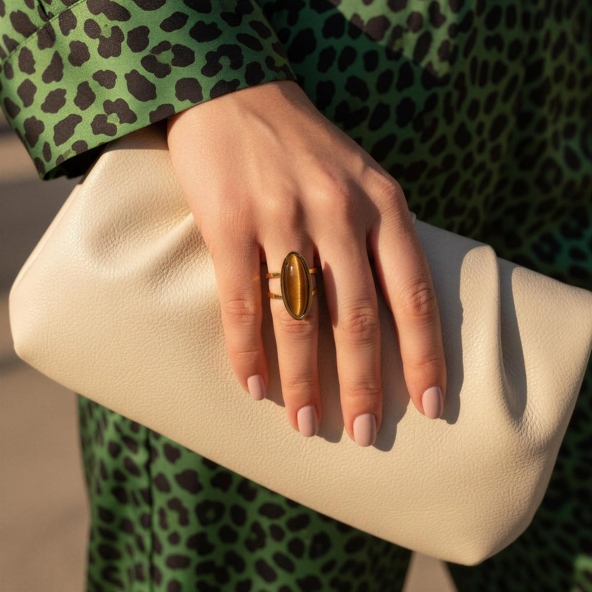 A close-up lifestyle shot of a person wearing the Sahara Ring from Chica Chica Jewelry,  a chunky oval gold ring with a brown tiger's eye inlay, holding a cream-colored textured leather clutch bag. The person is wearing a green leopard print top.