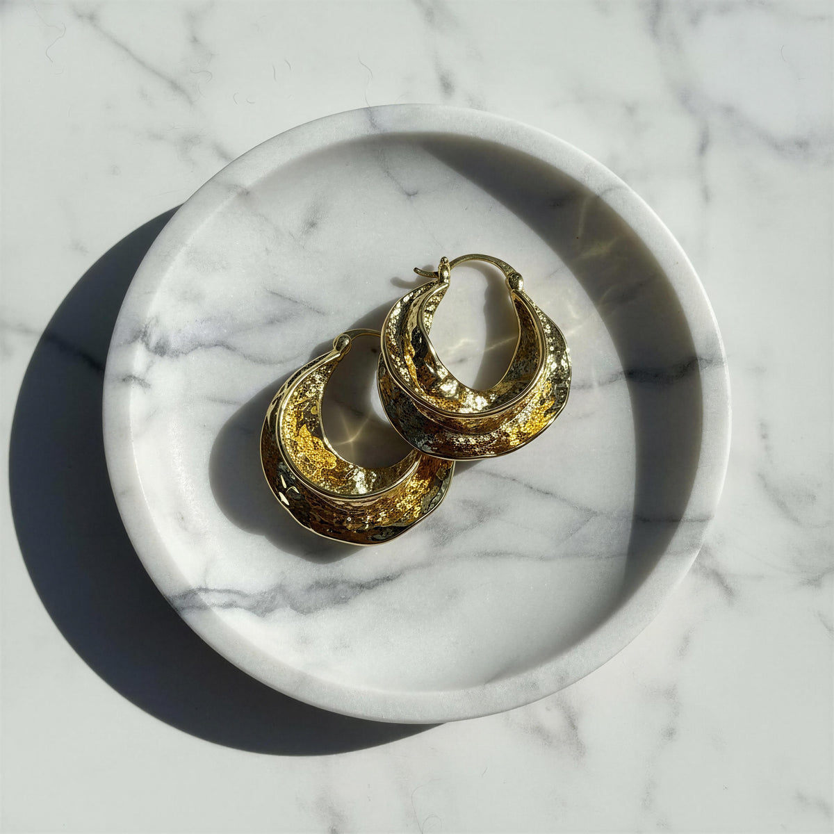 A pair of textured gold crescent-shaped hoop earrings resting on a white marble tray, set against a white marble surface with a hard shadow.