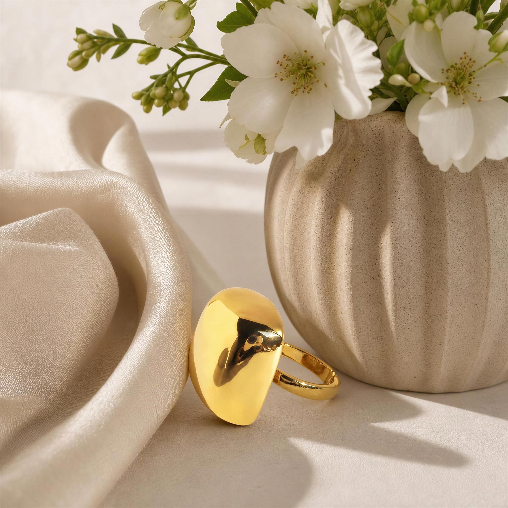  The Aurelia Ring from Chica Chica Jewelry resting on draped cream satin fabric next to a textured beige ceramic vase filled with white blossoms.