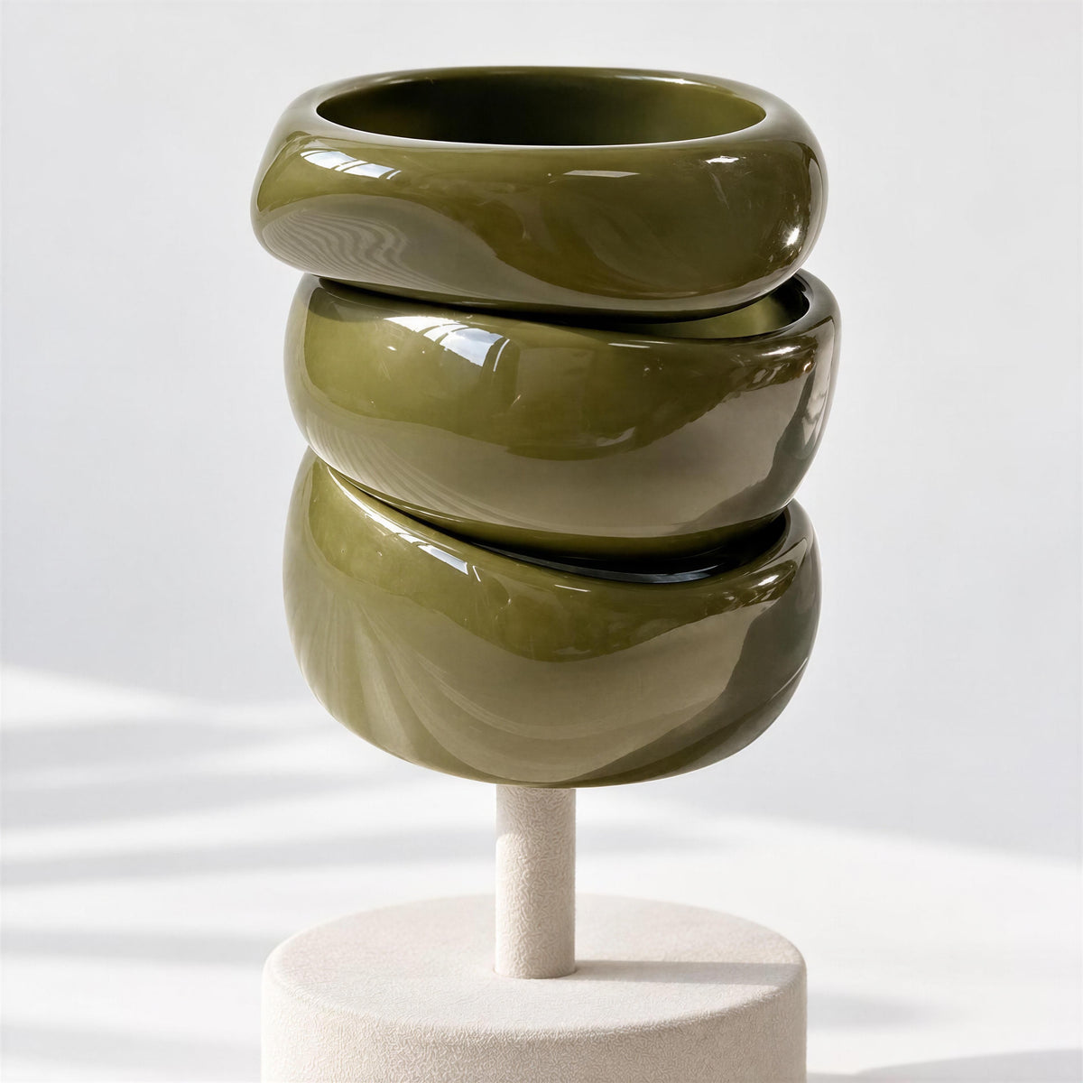The Oliva Bangle Set from Chica Chica Jewelry.   Three stacked glossy olive green ceramic bowls displayed on a light beige pedestal against a white background.