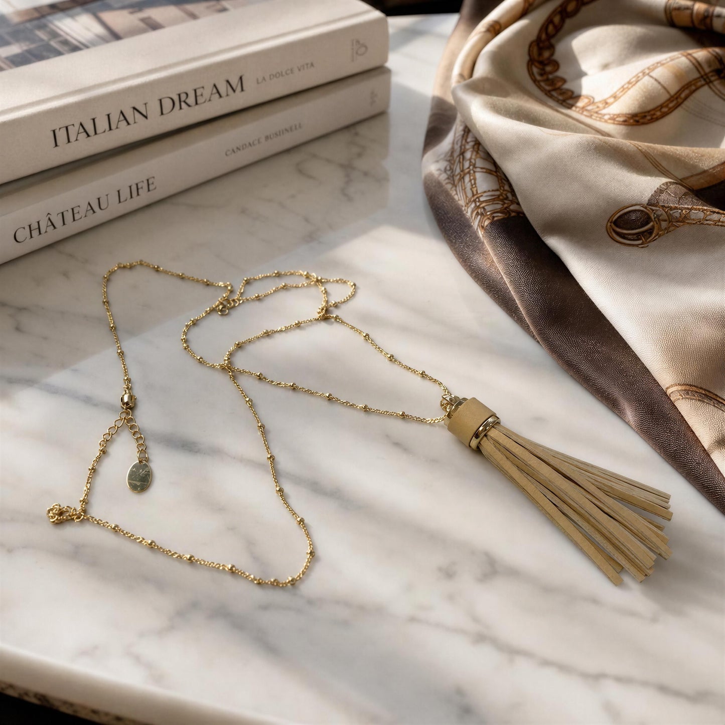 The Frangia Avorio Necklace, an 18k gold-plated  beaded chain necklace with a beige leather tassel, styled on a white marble surface next to books and a patterned scarf.