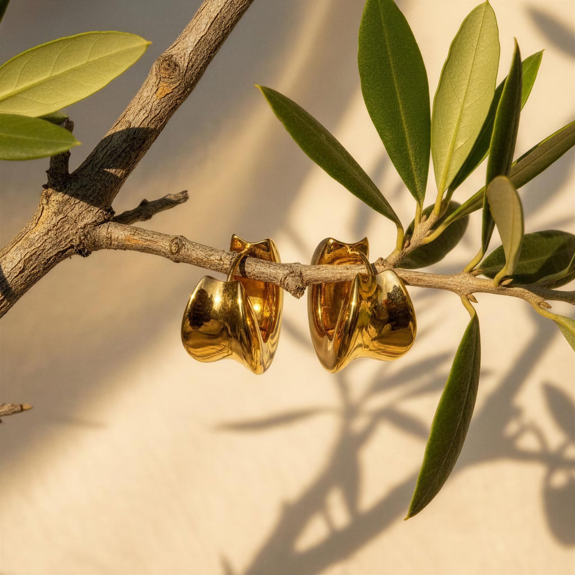 A pair of chunky, sculptural gold hoop earrings with a wavy, organic shape, hanging from a tree branch with green leaves in warm sunlight.