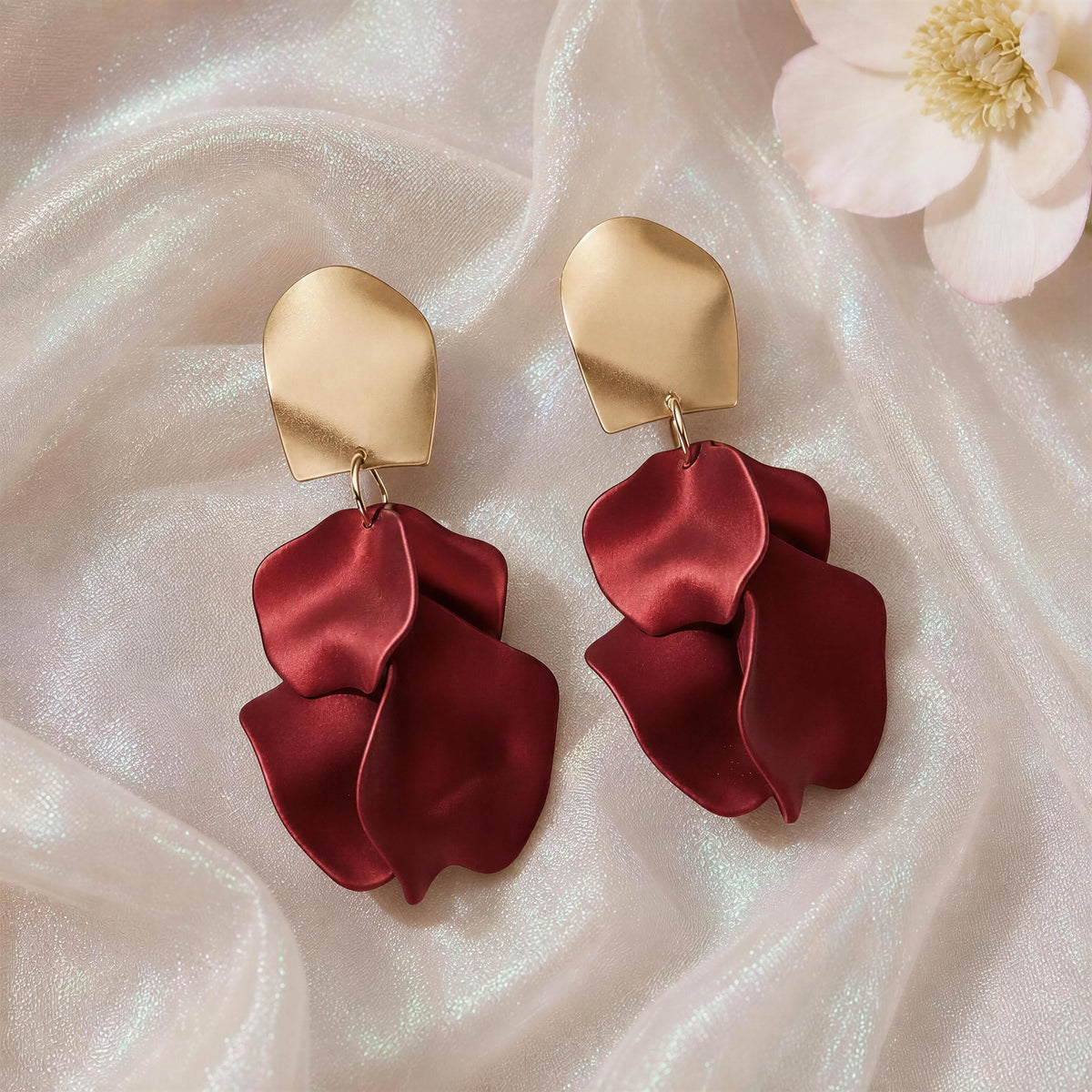 The Fiore Roja Earrings from Chica Chica Earrings.   A pair of statement dangle earrings with textured gold-tone top plates and layered deep burgundy satin petal drops, displayed on draped white fabric with a flower prop.