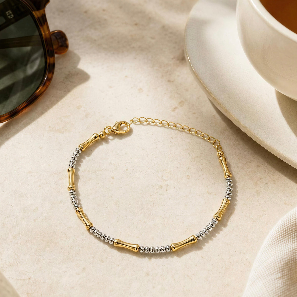 A delicate gold chain bracelet with alternating polished gold tube links and textured silver bead sections, displayed on a light beige stone surface next to a white cup and sunglasses.