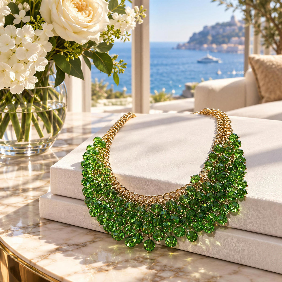 The Eleonora Necklace from  Chica Chica Jewelry.  A statement bib necklace made of green faceted gemstones and gold-toned metal, displayed on stacked white books on a marble table with a vase of white flowers and a luxurious seaside terrace in the background.
