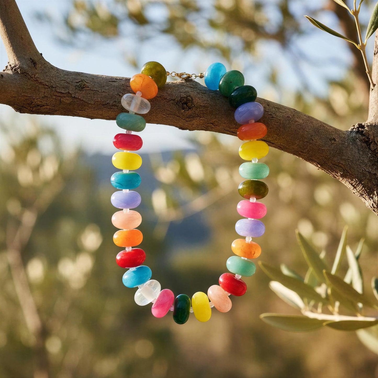 The Capri Multicolore Choker, a colorful beaded necklace made of round, disc-shaped beads in rainbow colors, hanging from a tree branch outdoors with a blurred natural background.