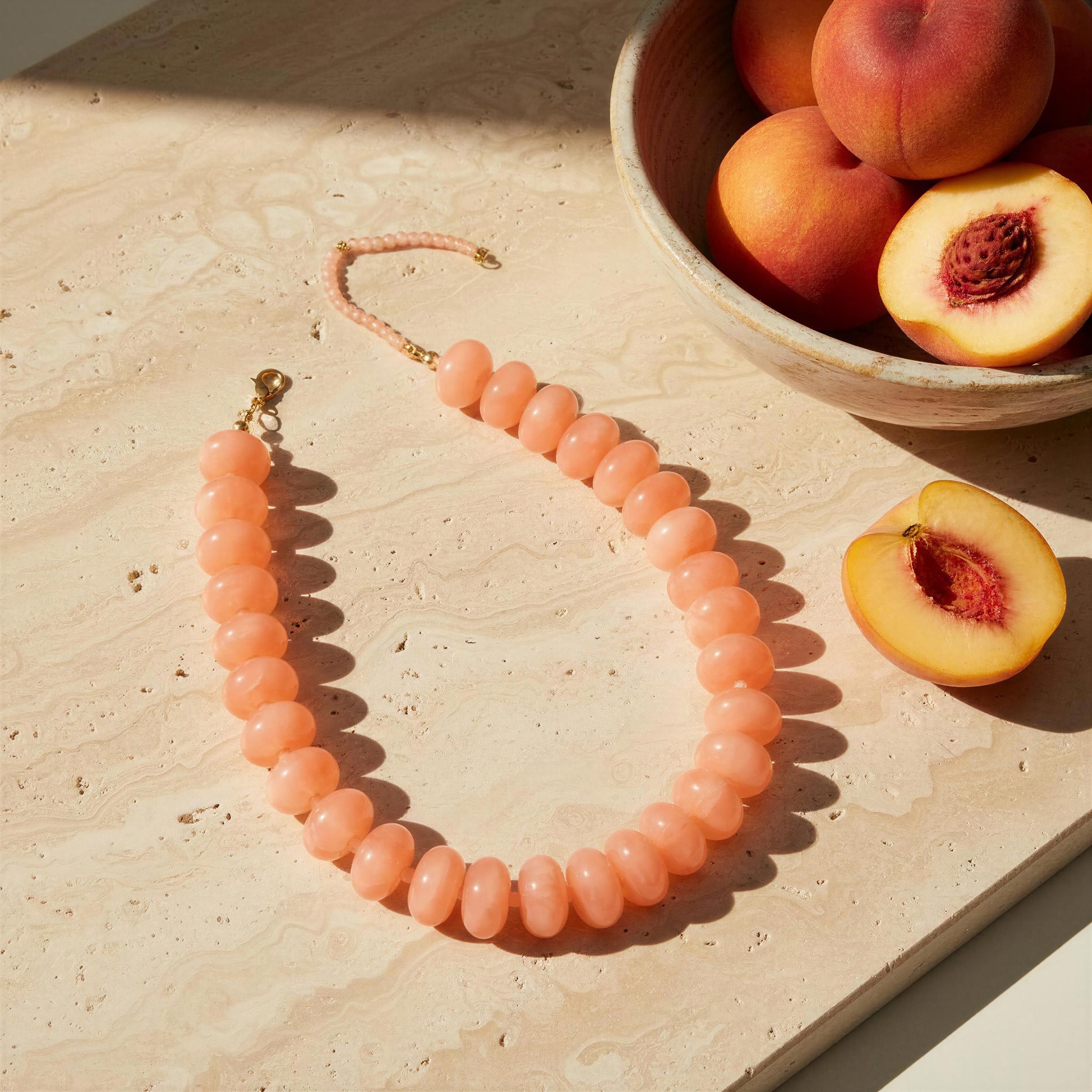 A peach-colored beaded necklace with gold-toned hardware lies on a light stone surface next to a bowl of peaches and a sliced peach in warm sunlight.