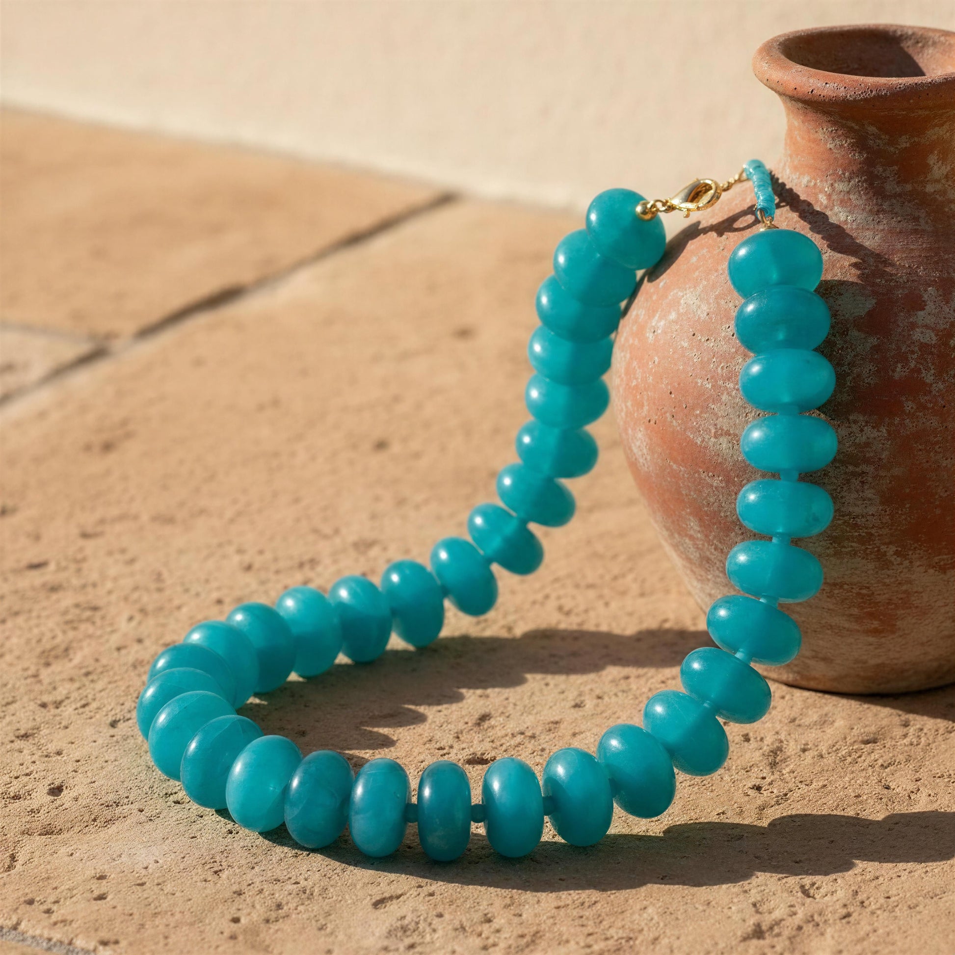 Turquoise beaded necklace on a sandy surface with a terracotta pot.
