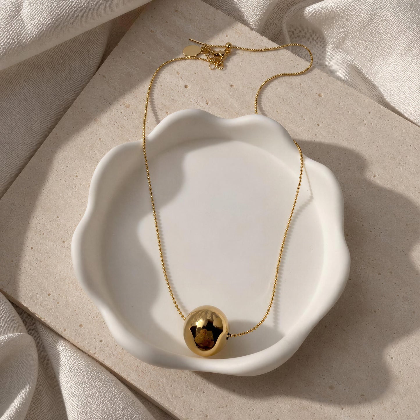 The Riviera Orb Necklace on a white dish against a beige fabric background 