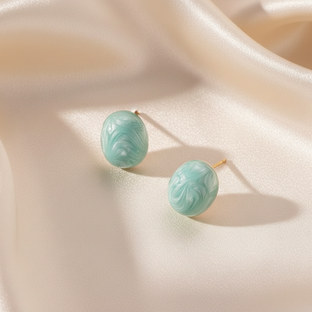Aqua marbled oval stud earrings displayed on soft ivory fabric, polished statement studs with ocean-inspired color.