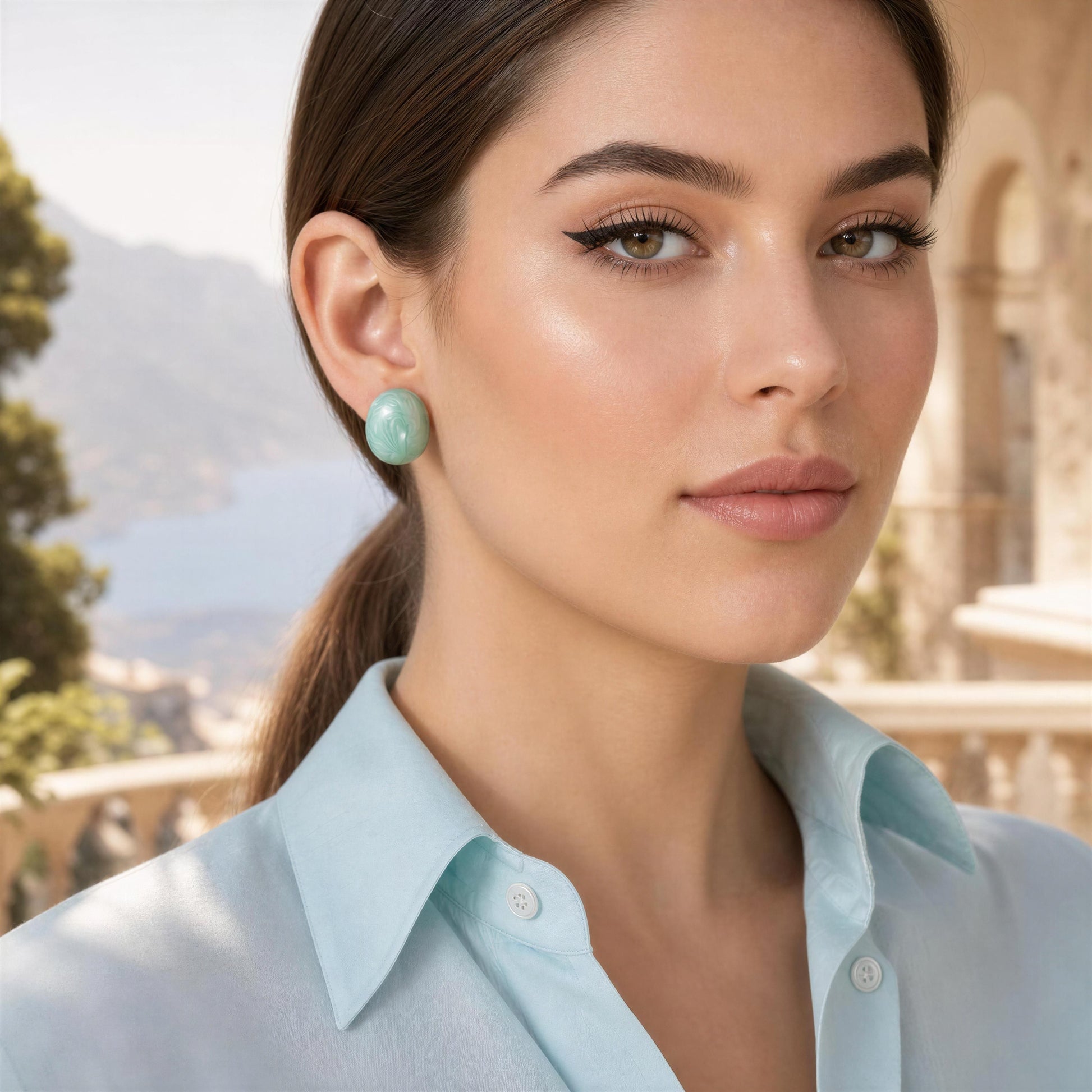 Woman wearing light blue shirt and the Rosina Studs in Aqua Glow with blurred outdoor background