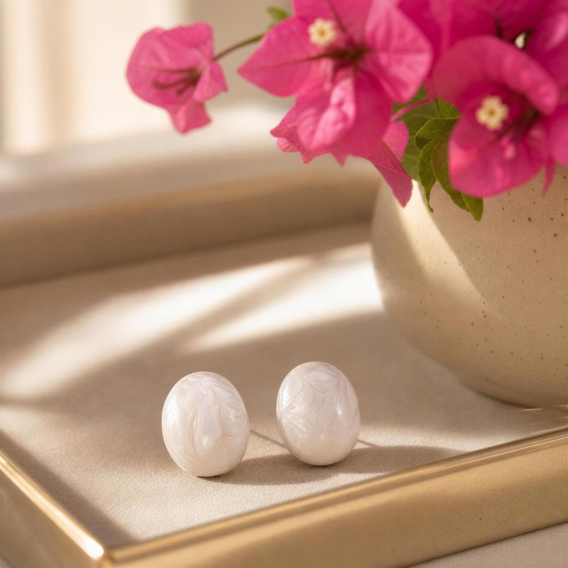 White marble-like earrings on a reflective surface with pink flowers in the background