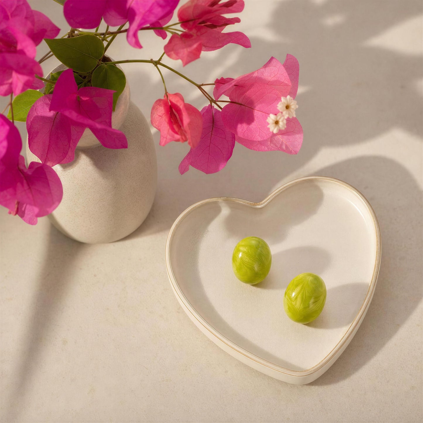 Heart-shaped dish with the  Rosina Studs - Verde Luxe on a beige surface next to pink flowers in a vase.