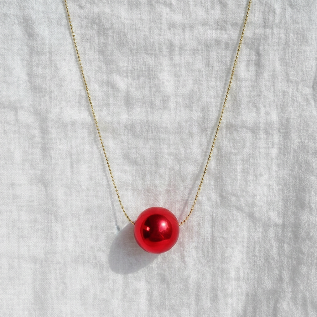 The  Rubi Petite Sphere Necklace with a red spherical pendant on a white fabric background