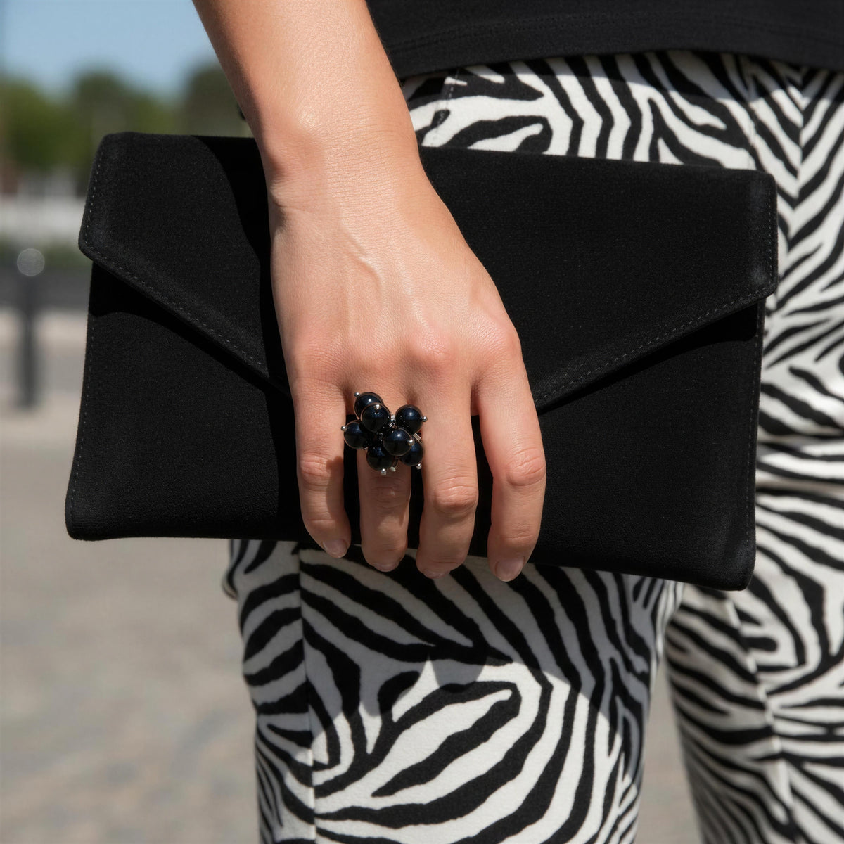 A person holding a black envelope-style clutch bag, wearing black and white zebra-print pants and a large black beaded ring on one finger, photographed outdoors.