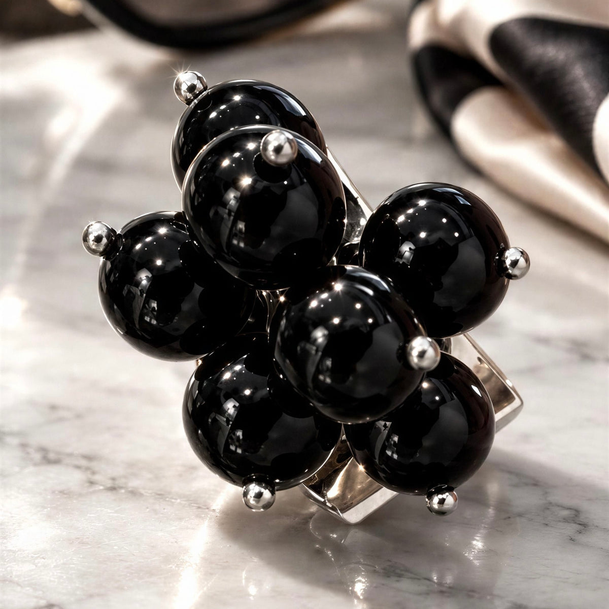 The Sabina Ring from Chica Chica Jewelry - a statement ring featuring seven glossy black onyx-like beads clustered on a silver-toned band, photographed on a light marble surface with a blurred black-and-white fabric in the background.