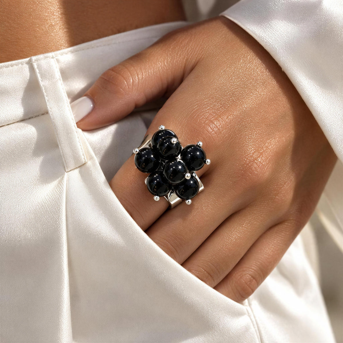 A close-up of a hand wearing the Sabina Ring  from Chica Chica Jewelry, a large silver ring with seven glossy black beads, resting on white satin fabric.