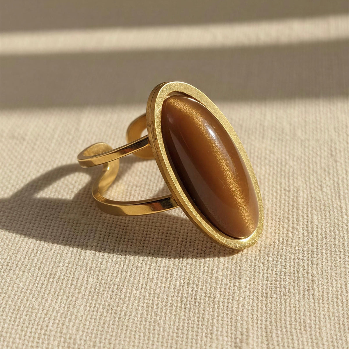 The Sahara Ring from Chica Chica Jewelry. A gold ring with a large, polished oval brown cat-eye stone, resting on a textured beige fabric under warm, direct sunlight.