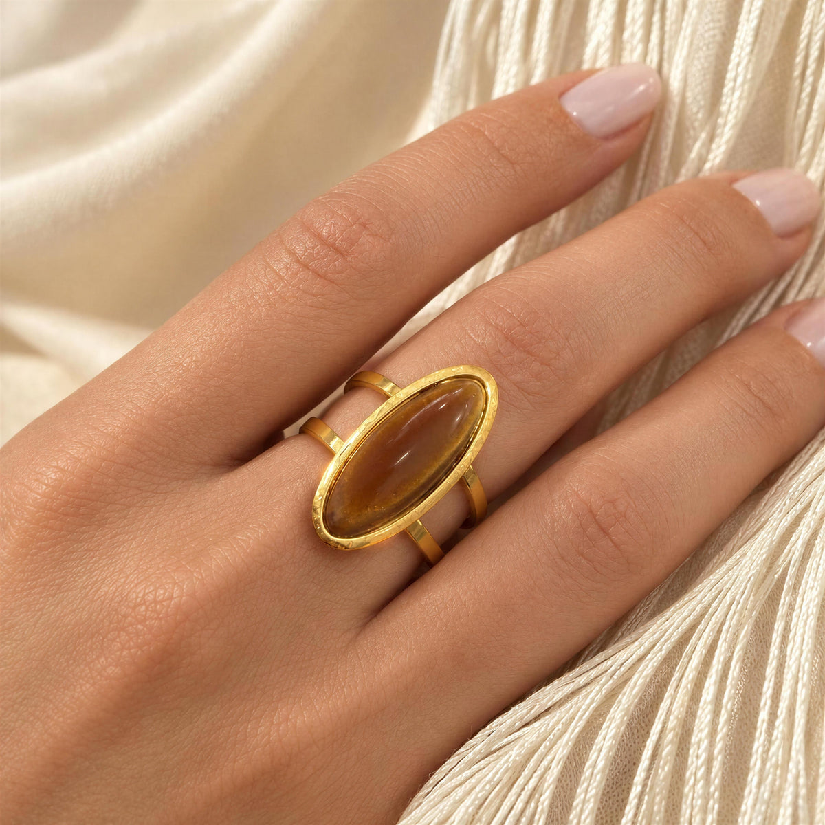 The Sahara Ring from Chica Chica Jewelry.  A close-up of a hand wearing a gold ring with a large oval brown cabochon stone, resting against draped cream-colored fabric.