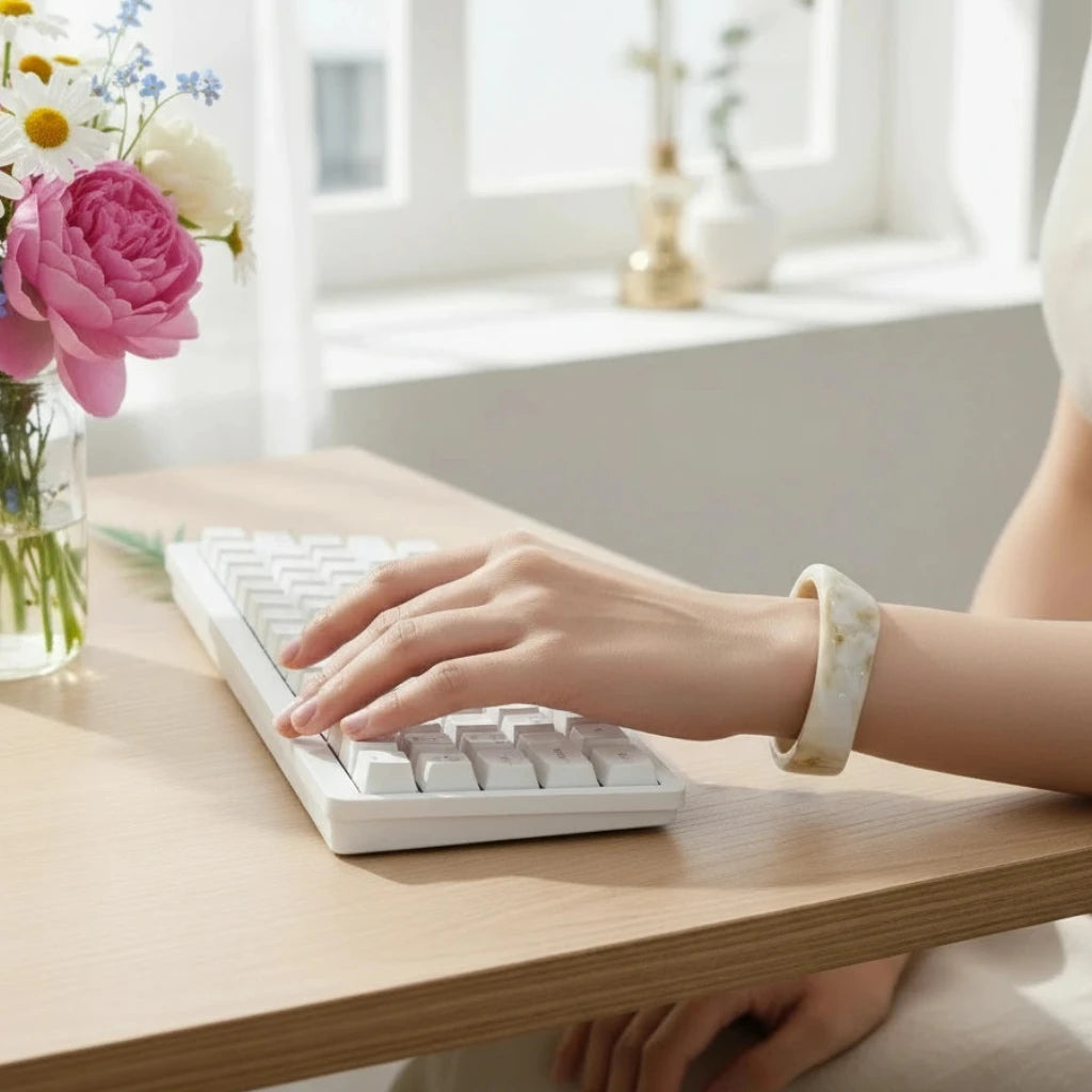 Person typing on a white keyboard with a vase of flowers on a wooden desk and the Sandy Glow Bracelet
