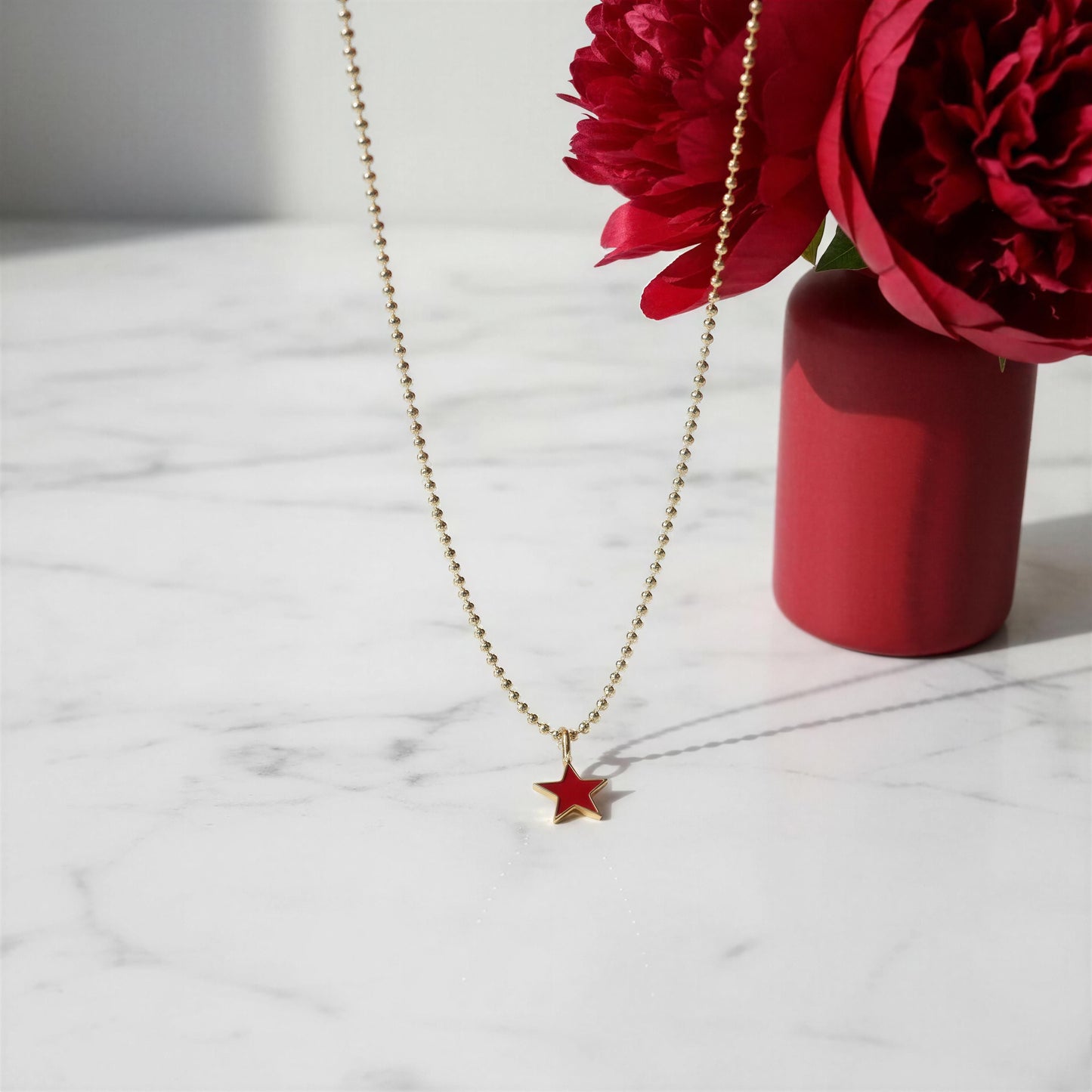 The Scarlet Star Necklace from Chica Chica Jewelry on a marble surface with red flowers.