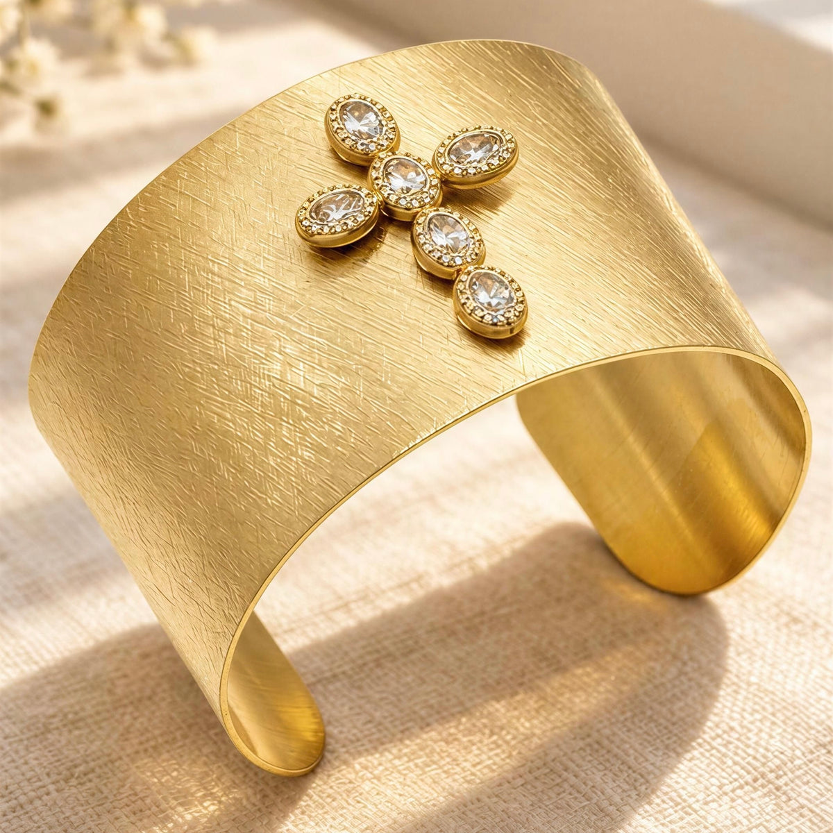 The Serafina Cuff from Chica Chica Jewelry. A wide gold cuff bracelet with a brushed finish and a decorative cluster of five clear oval gemstones at the front, photographed on a light beige textured surface.